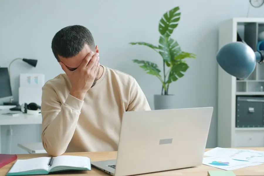 A man in a beige sweater sits at a wooden desk with one hand covering his eyes, an open silver laptop in front of him, an open notebook and loose papers on the desk, and a potted plant and white shelving behind him in a bright room.