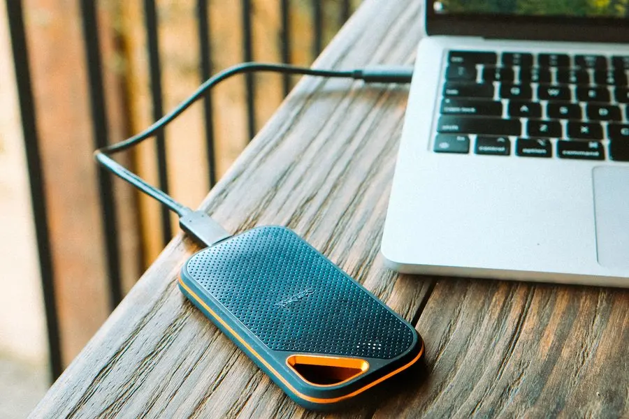 A dark grey portable external drive with orange trim resting on a weathered wood surface, connected by a black USB-C cable to an open silver laptop beside it.