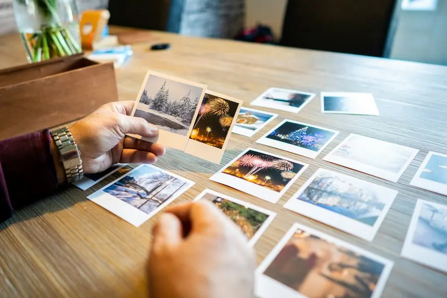 A person's hands sorting dozens of printed colour photographs spread across a light wood table, holding two of them up, with an open wooden keepsake box at the left edge.