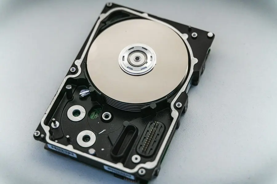 An opened hard drive lying on a pale grey surface with its top cover removed, showing the mirrored disc platter, the central spindle hub, and the screws and ribbon connector around the black metal frame.