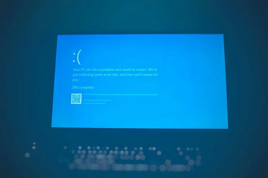 A laptop screen glowing in a dim room, displaying a blue error page with a sad-face emoticon and text reading that the PC ran into a problem and needs to restart while it collects error info, marked 20 percent complete, with the backlit keyboard blurred below.