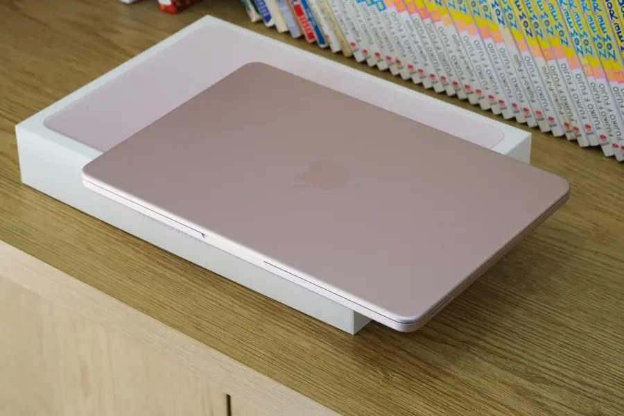 A closed pale-pink laptop resting on top of its opened white retail box on a light wooden cabinet, with a row of colourful book spines on a shelf behind it.