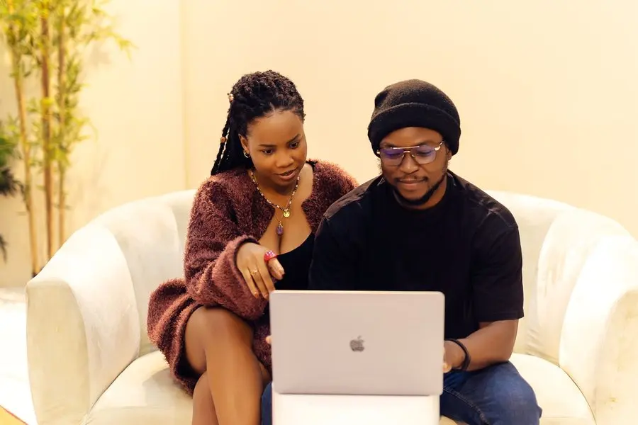 A woman in a brown fuzzy cardigan and a man in a black beanie and glasses sit side by side on a cream sofa, both leaning in and looking down at an open silver laptop.