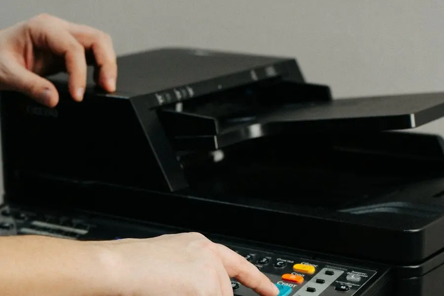 Two hands at a black all-in-one printer: one lifts the document feeder lid, the other presses a button on the control panel.