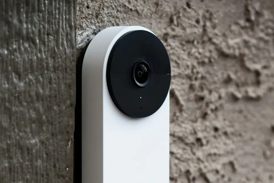 A tall white video doorbell with a dark circular camera lens and a small green indicator light, mounted on wooden door trim against a textured stucco exterior wall.
