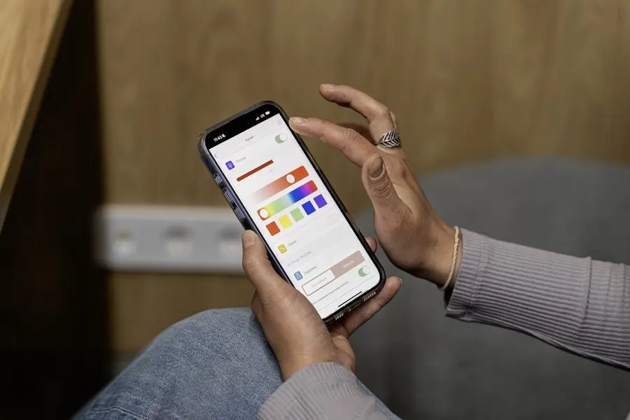 A seated person in a grey sweater and jeans holds a smartphone in both hands, thumb reaching toward a lighting control screen showing a brightness slider and a row of colour swatches, with a wood-panelled wall and a grey upholstered chair behind.