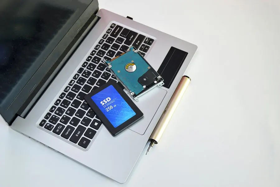 A black 2.5-inch solid-state drive with a blue label reading SSD 256 GB, lying on the keyboard of an open silver laptop next to an older bare spinning hard drive mounted in a metal caddy, with a gold pen beside them on a white desk.