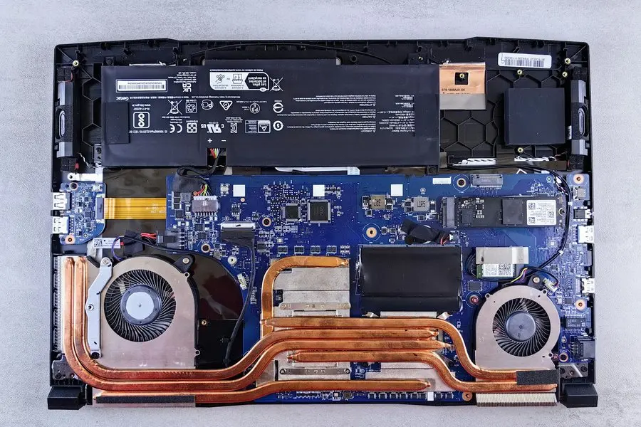 The underside of a laptop with its bottom cover removed, seen from directly above: a flat black battery across the top, a blue circuit board, two cooling fans joined by copper heat pipes, and a small M.2 solid-state drive seated in a slot on the right beside an empty second slot.