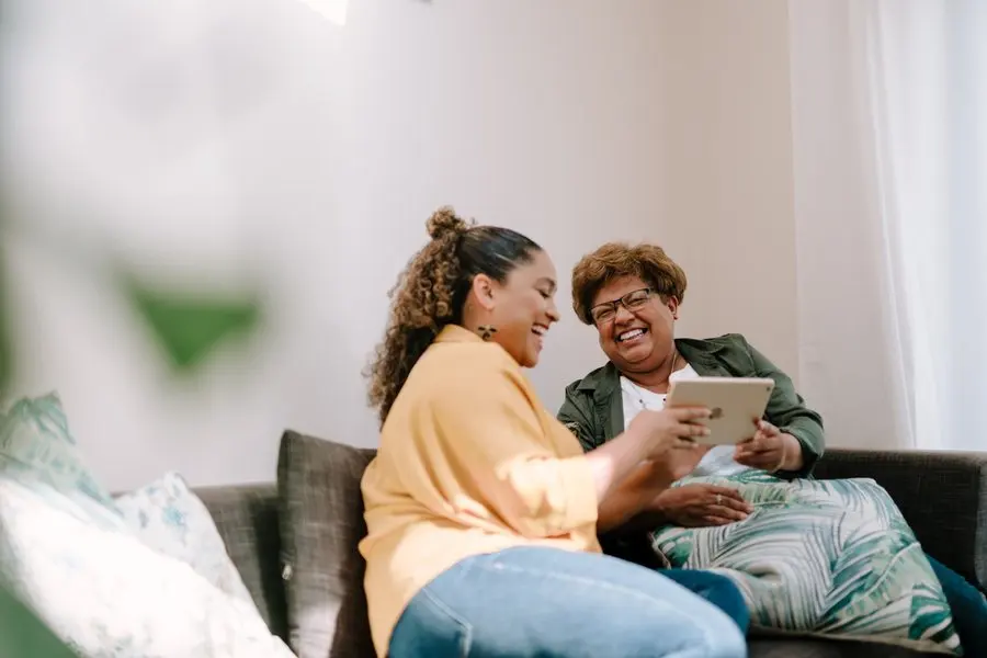 Two women sit together on a grey couch in a bright living room, laughing. The younger woman, in a yellow shirt and jeans, holds a white tablet turned toward the older woman, who wears glasses and a green jacket and is looking at the screen.
