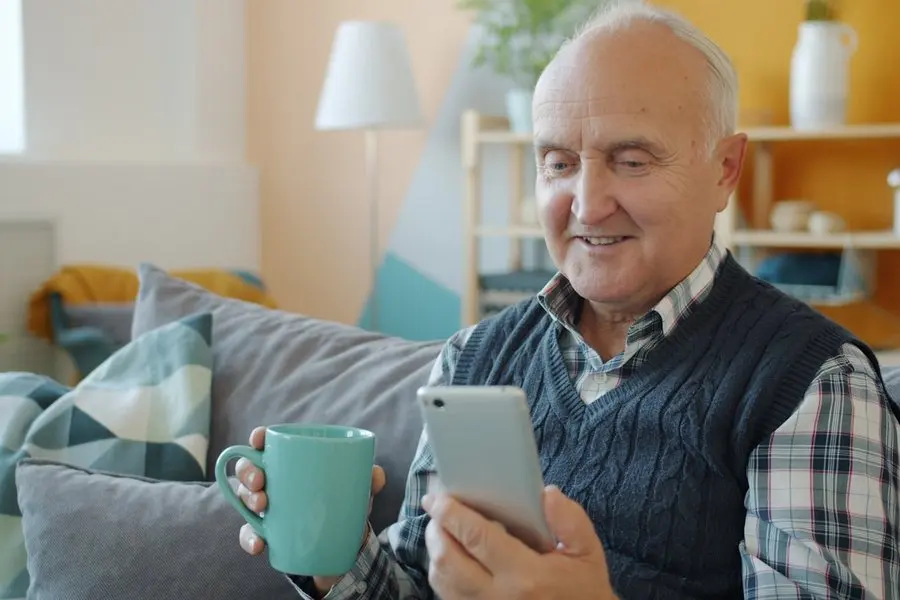 An older man with white hair, wearing a plaid shirt under a navy knit vest, sits on a couch smiling at a silver smartphone he holds in one hand while holding a mint-green mug in the other.