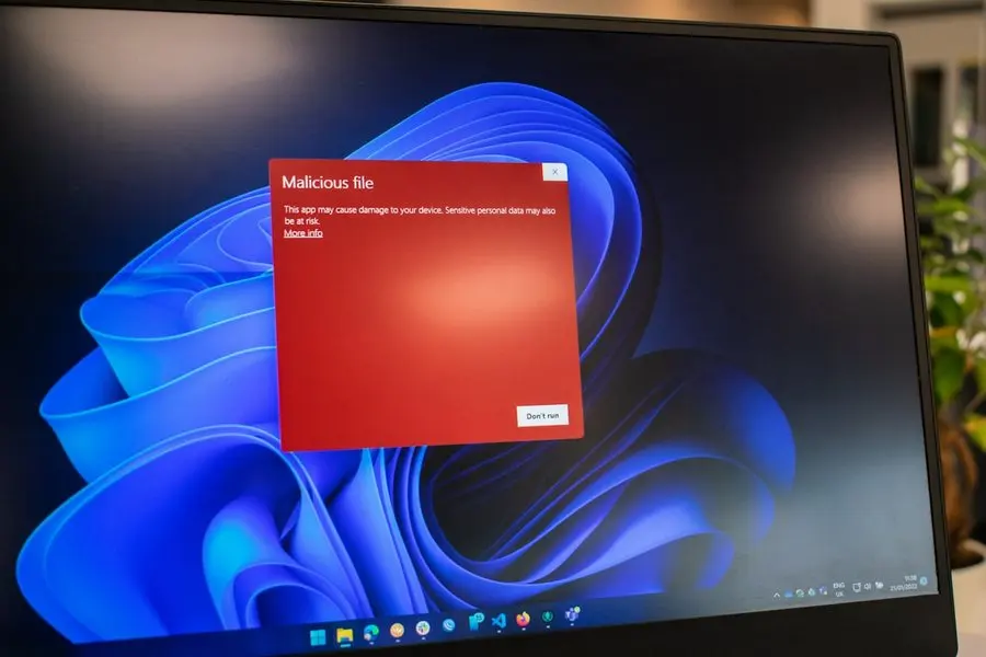 A laptop screen showing a red pop-up warning box titled "Malicious file" over a blue desktop background. The message reads that the app may cause damage to the device and that sensitive personal data may also be at risk, with a "More info" link and a "Don't run" button.