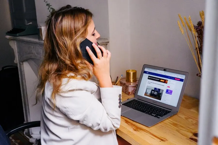A woman sits at a wooden desk in a home, holding a mobile phone to her ear with one hand while looking at the screen of an open laptop in front of her. A stone fireplace mantel and a vase of dried grasses are behind her.