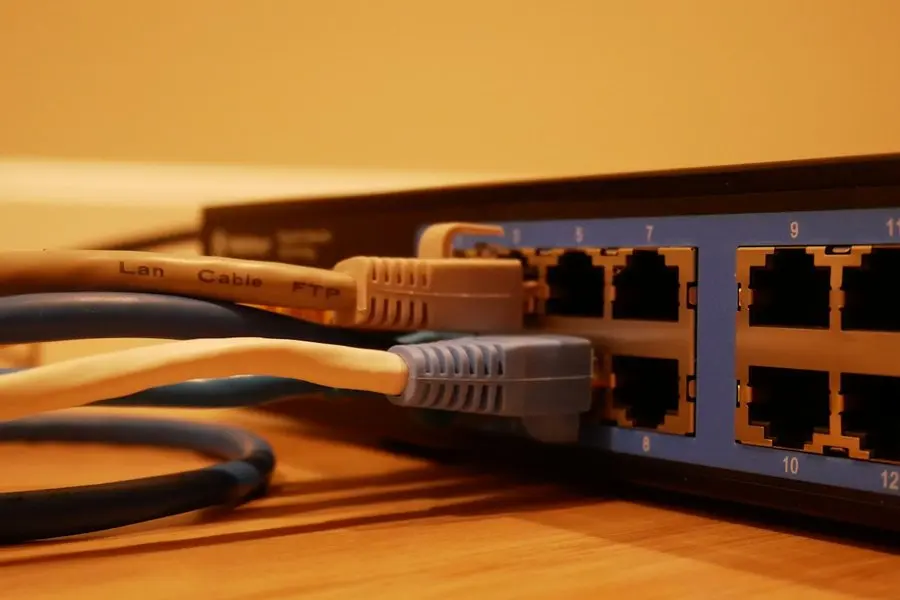 Close-up of a black multi-port network switch on a wooden surface, its numbered port bank filled with beige and pale-blue ethernet cables under warm indoor light.