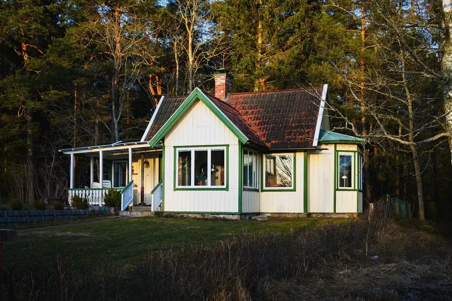A small white cottage with green window trim and a tiled roof standing on its own in a grassy clearing surrounded by tall pine trees, lit by low evening sun.